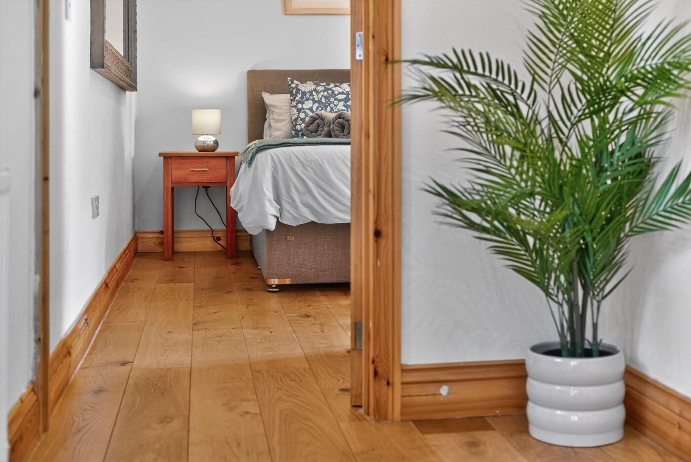 A narrow hallway with a wooden floor, a bed with a headboard, a bedside table with a lamp, and a potted plant.
