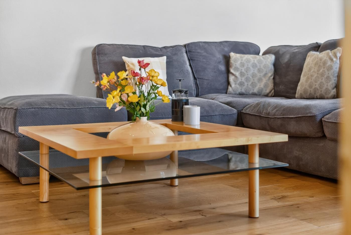 A modern living room with a glass-top coffee table, a vase with flowers, and a grey sofa with patterned cushions.