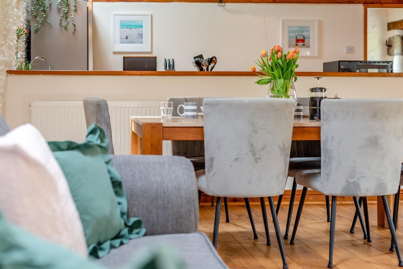 Un comedor con una mesa de madera, dos sillas grises, un jarrón con flores y una zona de cocina con electrodomésticos al fondo.