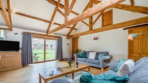 Een ruime woonkamer met een houten plafond, een grijze zithoek, een salontafel en een televisie op een houten kast.