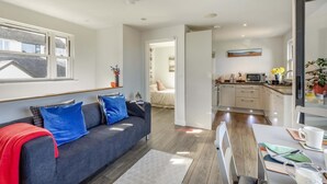 Uma sala de estar moderna com sofá, área de jantar com mesa e cadeiras, cozinha com armários e eletrodomésticos, e corredor que leva a outro quarto.