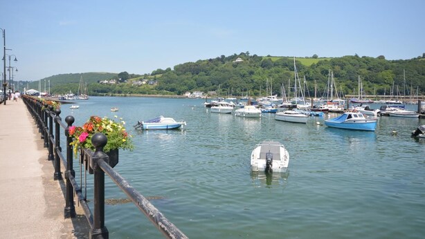 Uma marina com barcos atracados, um calçadão para pedestres e uma colina arborizada ao fundo.
