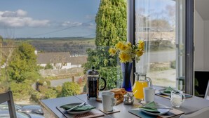 Uma mesa de jantar com vista para uma paisagem rural, uma cafeteira, um jarro de suco de laranja e um vaso com flores amarelas.