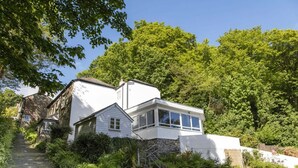 Une maison à deux étages avec un escalier en pierre, entourée d’une végétation luxuriante et d’arbres.