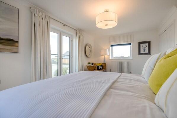 A hotel room with a bed, a window with curtains, a bedside table with a lamp, and a wall clock. - 4 Bed in Padstow (oc-p36067) (Constantine Bay)