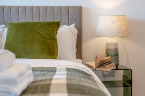 Ein ordentlich bezogenes Bett mit einem grünen Kissen, einem weißen Kissen und einer karierten Decke. Neben dem Bett steht ein Nachttisch mit einer Lampe, einem offenen Buch und einer Steckdose.