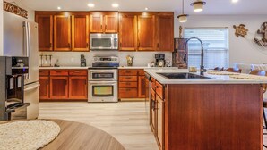Een keuken met houten kastjes, roestvrijstalen apparatuur, een groot eiland en uitzicht op de woonkamer.