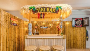 Een tiki bar met rieten dak, drie rieten barstoelen en een dartbord aan de muur.