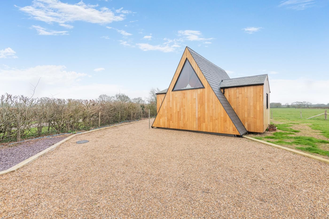 Un edificio moderno de madera con techo triangular y un gran ventanal, ubicado en un camino de grava con un campo de césped al fondo.