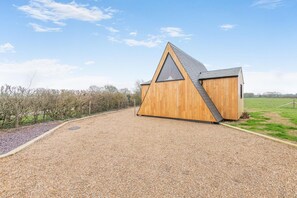 A modern wooden building with a triangular roof and a large window, situated on a gravel driveway with a grassy field in the background.