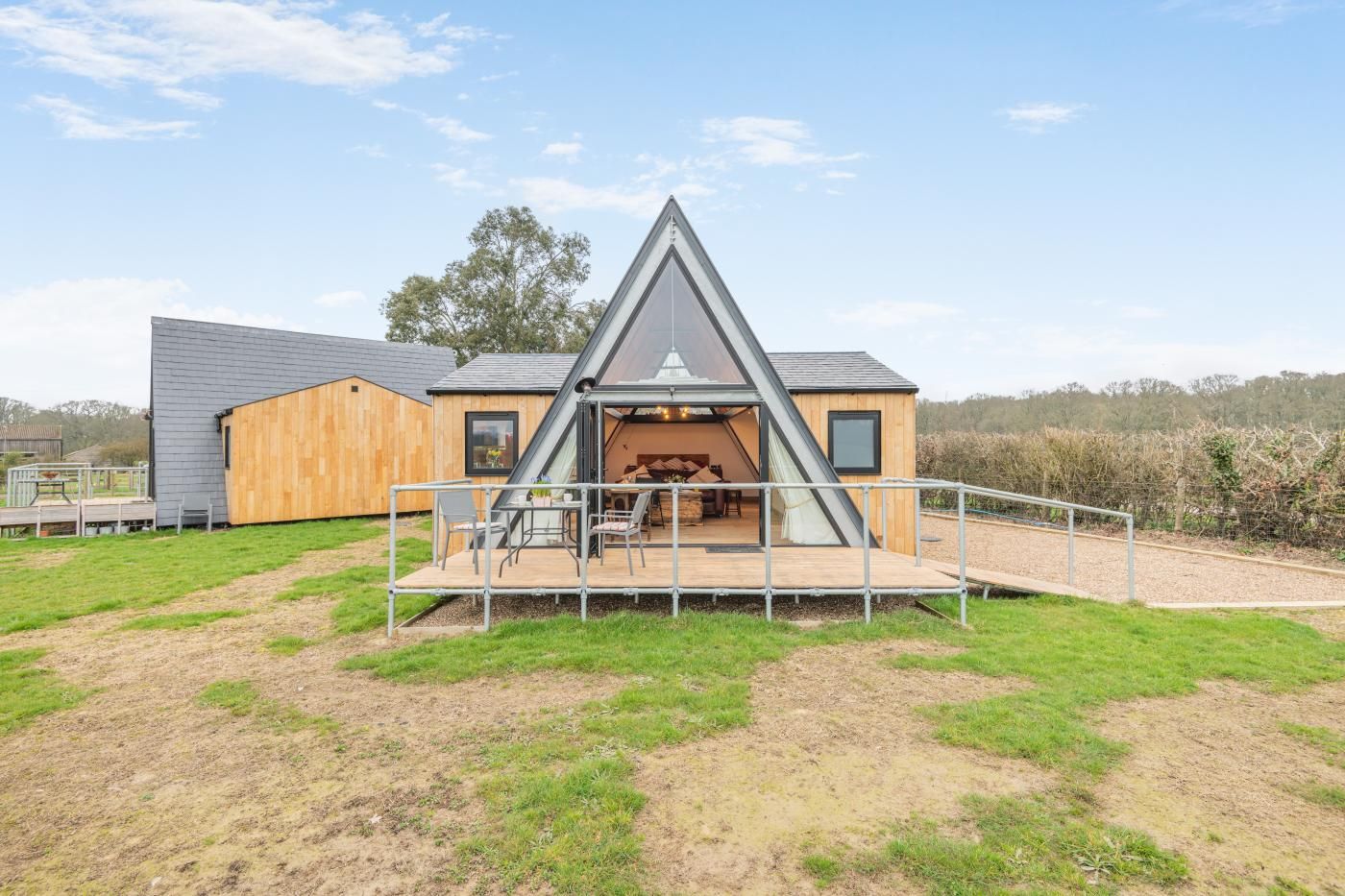 Una casa moderna con techo triangular, una terraza de madera y un área exterior cercada.