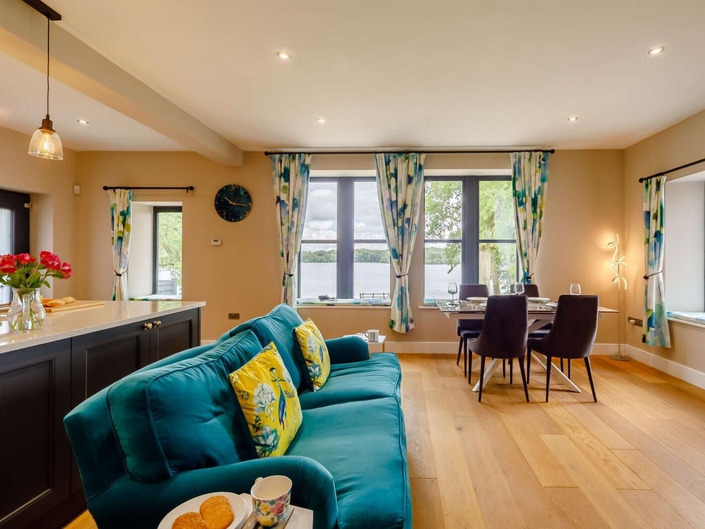 A modern living room with a teal sofa, a dining area with a table and chairs, a kitchen area with a counter, and large windows with curtains.