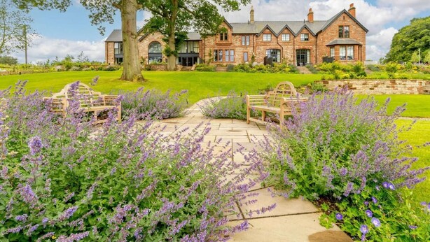 A large brick house with multiple windows, a lawn, and a garden with purple flowers. - 2 Bed in Brampton (oc-88663) (Brampton)