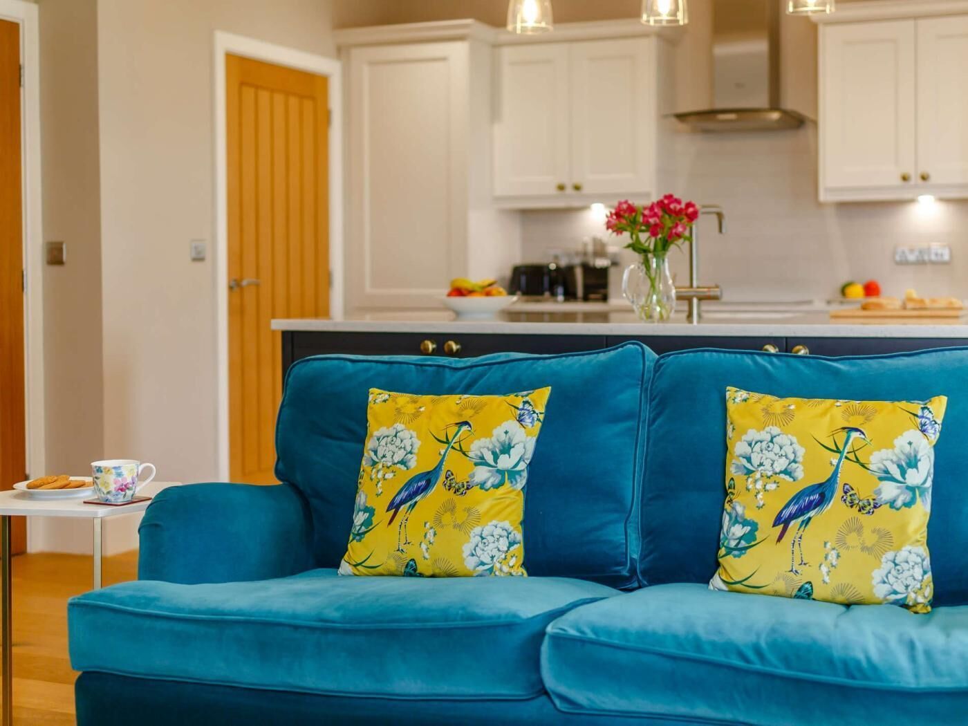A blue sofa with floral cushions, a white table with a cup and plate, and a kitchen in the background.