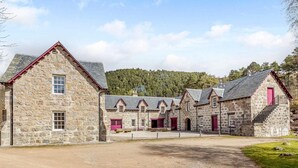Un bâtiment en pierre comprenant plusieurs pièces, une allée gravillonnée et, au loin, une colline boisée.