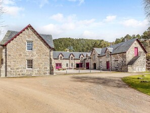A stone building with multiple rooms, a gravel driveway, and a forested hill in the background. - 10 Bed in Alness (oc-ca233) (Alness)