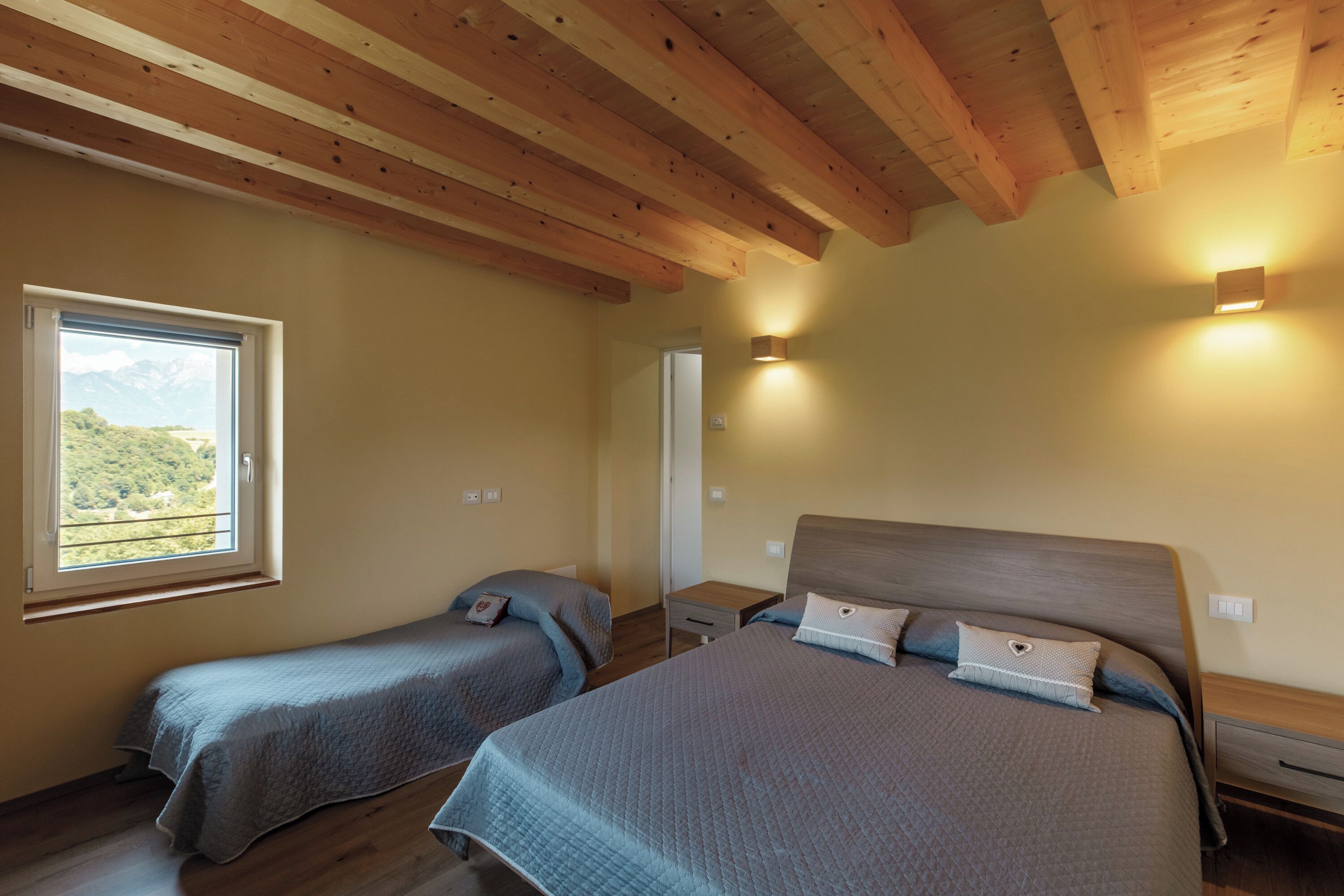 A bedroom with two beds, wooden beams on the ceiling, a window with a view, and bedside lamps.