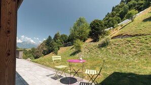 A patio with a table and chairs, a wooden post, and a view of a grassy hill with trees and a mountain in the background.