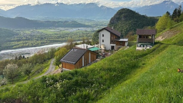 A hillside property with a modern house, a wooden cabin, and a view of a river and mountains.