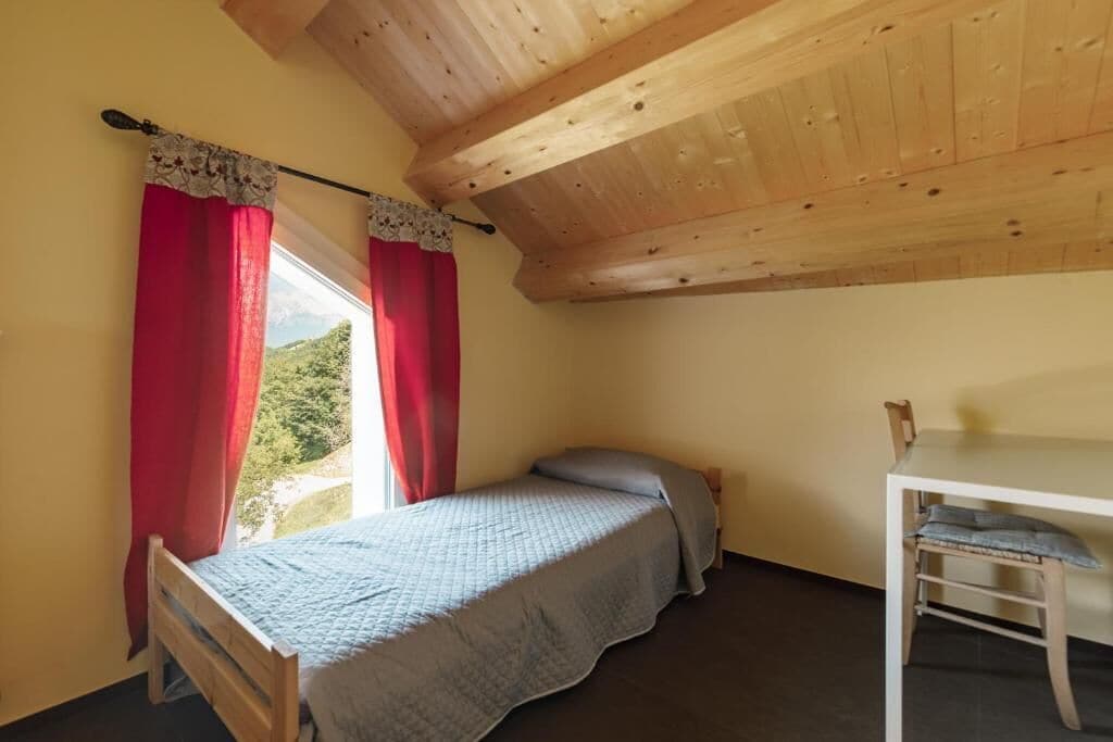 Une petite chambre avec un lit simple, un plafond en bois, une fenêtre avec des rideaux rouges et une petite table avec une chaise.