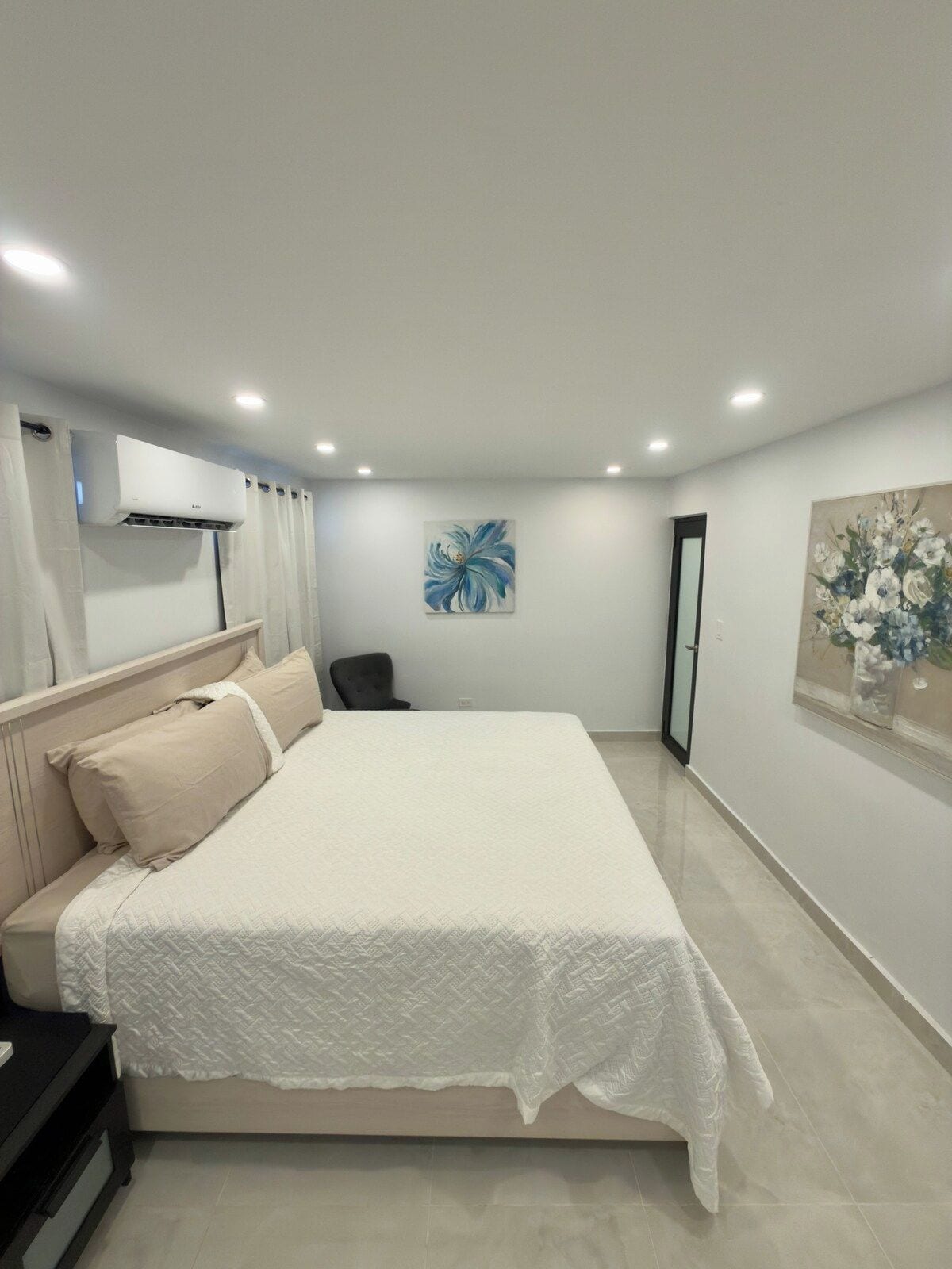 A bedroom with a bed, a chair, an air conditioning unit, and a painting on the wall.