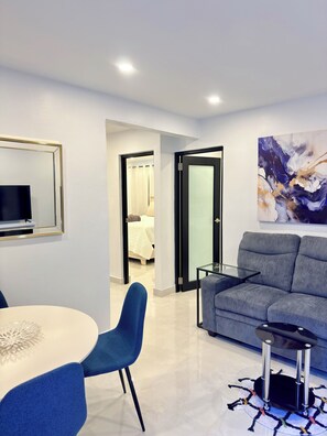 Ein modernes Wohnzimmer mit grauem Sofa, einem Glastisch, einem blauen Sessel, einem an der Wand montierten Fernseher und einem Gemälde.