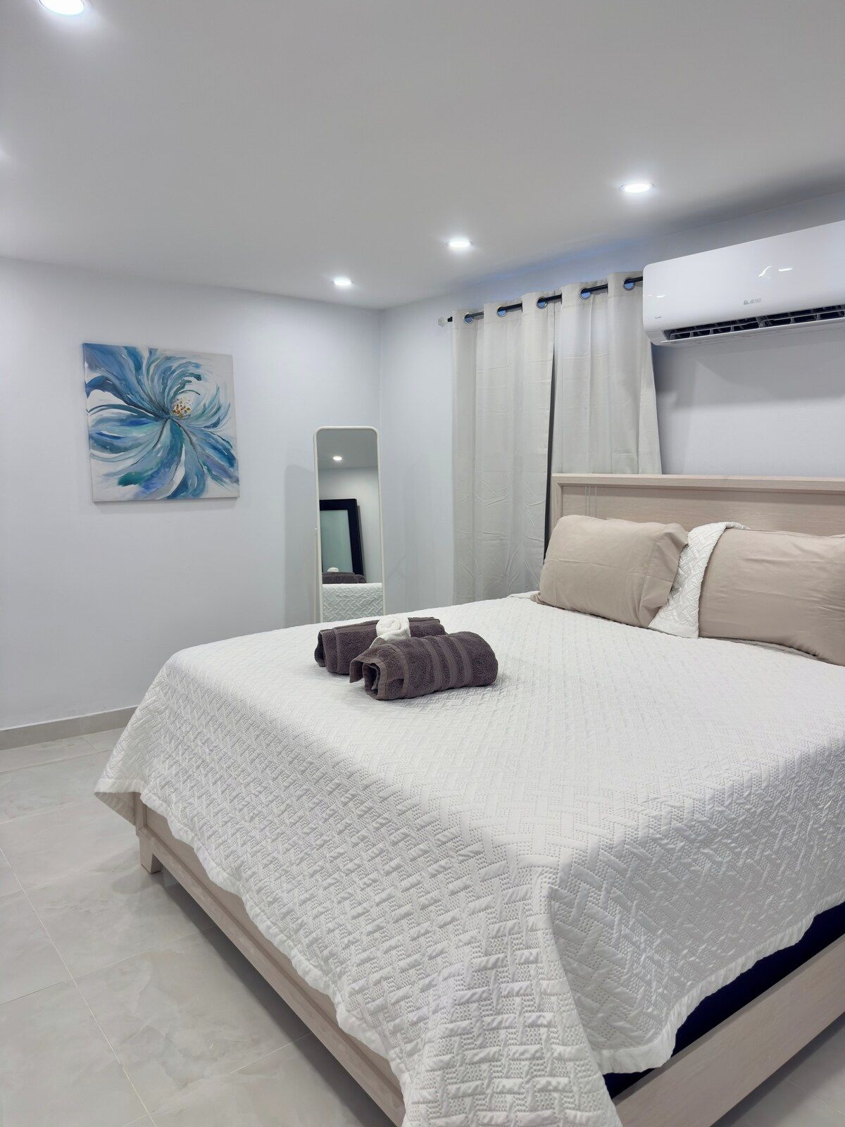 Un lit bien fait, recouvert d’une couette blanche et agrémenté de deux serviettes enroulées, un grand miroir au mur et un appareil de climatisation au-dessus du lit.