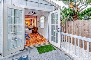 A house with a wooden deck, a sliding glass door leading to a room with a ceiling fan, and a small outdoor area with a green mat. - Azul 's House - Your Tiki Bar Getaway, Game Room, and Seashell-Filled Backyard (Venice)