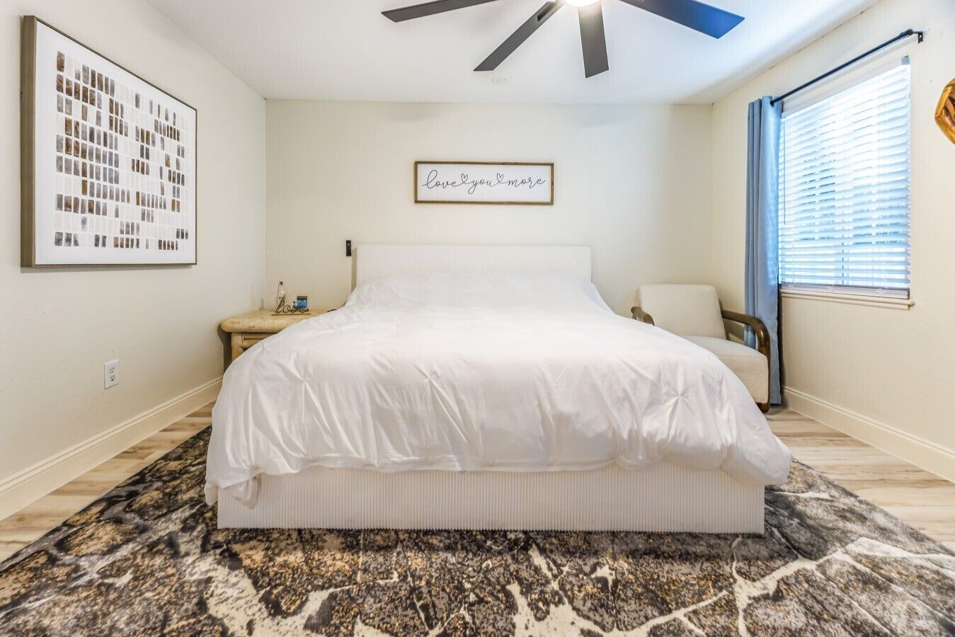 A bedroom with a large bed, a nightstand, a chair, a framed picture, and a ceiling fan.