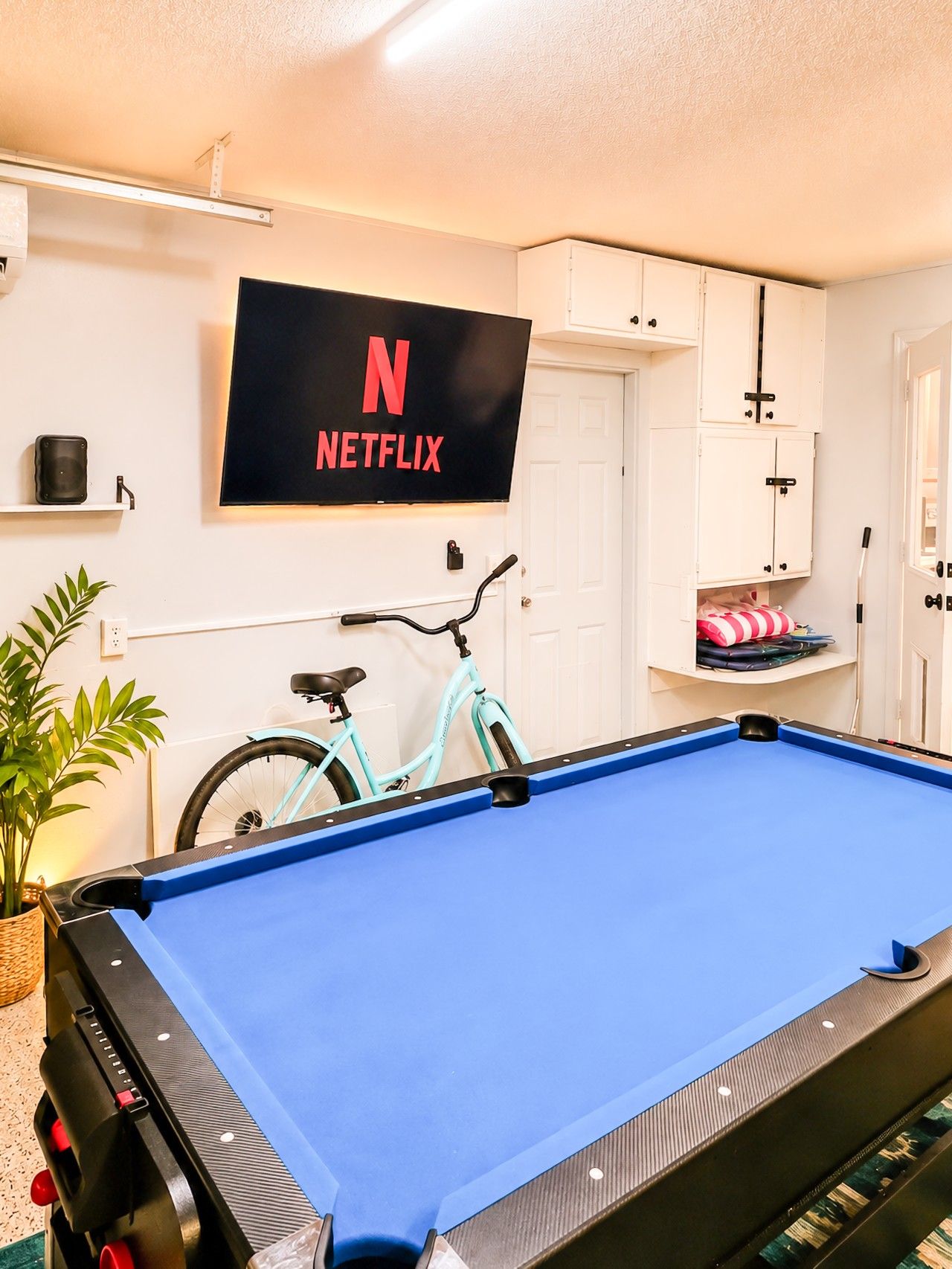 Uma mesa de bilhar em um aposento com uma televisão montada exibindo o logotipo da Netflix, uma bicicleta e armários.