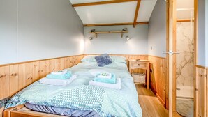 A room with a bed, wooden walls, a ceiling with wooden beams, a small bedside table, and a door leading to a bathroom.