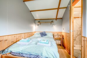 A room with a bed, wooden walls, a ceiling with wooden beams, a small bedside table, and a door leading to a bathroom.