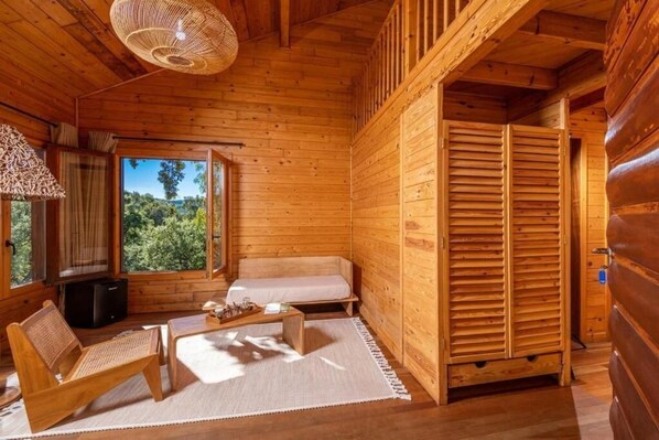 A wooden cabin interior with a sofa, a chair, a table, and a large window offering a view of greenery.