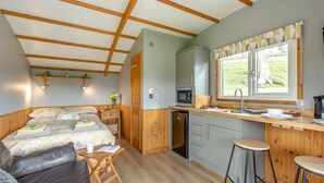 Een gezellige slaapkamer met een bed, nachtkastje en een klein houten krukje. Grenzend daar aan een compacte keuken met een spoelbak, fornuis en magnetron.