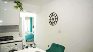 A compact kitchen with a washing machine, a dining table with a chair, and a clock on the wall.