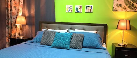 A bed with a blue quilt, a grey and blue pillow, and a headboard. There are two bedside lamps, a green wall, and an air conditioning unit above the bed.