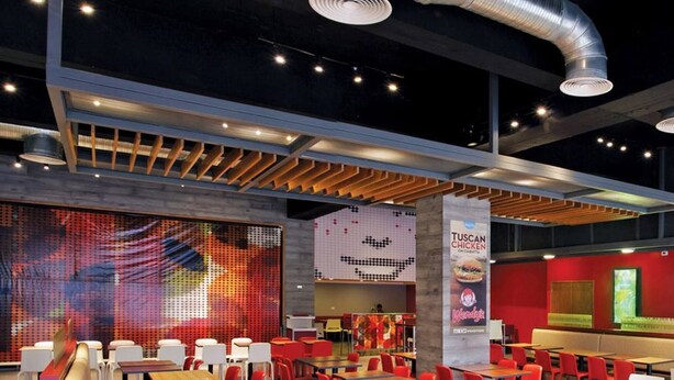 Interior moderno de restaurante com pé-direito alto, dutos expostos e paredes em tons de vermelho e neutros.
