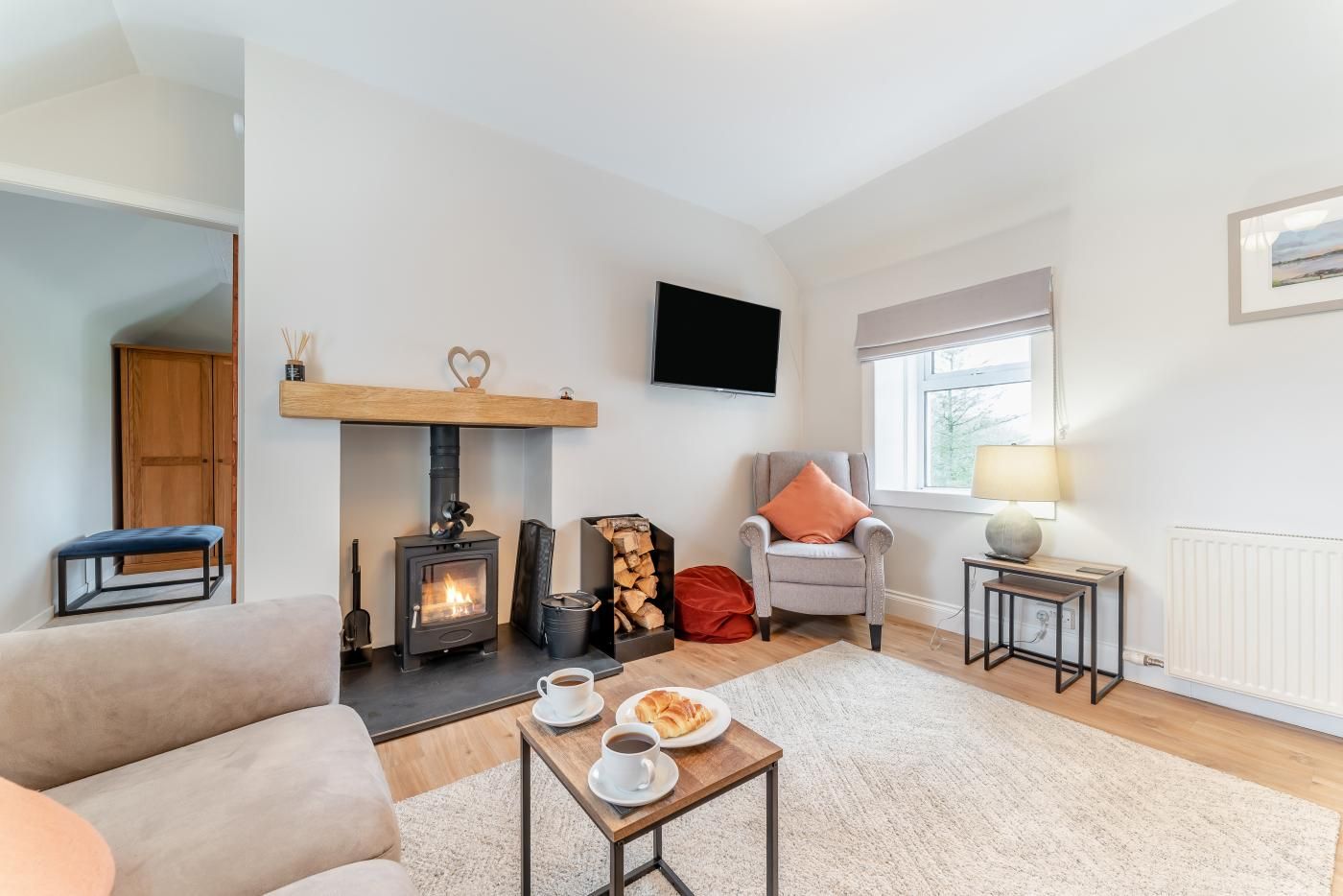 A cozy living room with a fireplace, a sofa, a coffee table with cups and a plate, a TV mounted on the wall, and a window with a view.