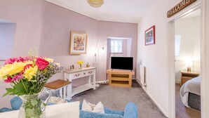 Ett rum med en säng, en tv på ett stativ, ett skrivbord med en stol, en vas med blommor och en skylt med texten ”VÄLKOMMEN”.