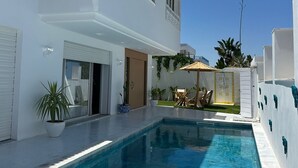 A modern white building with a swimming pool, a door, and a small patio area with a table and chairs.