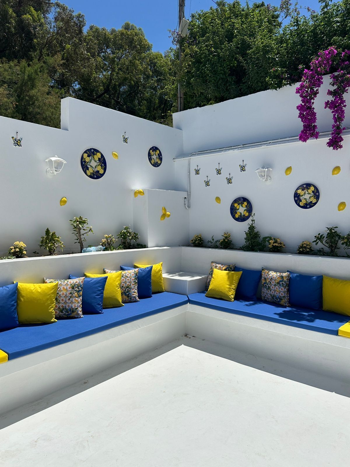 A white outdoor seating area with blue cushions and yellow pillows, surrounded by white walls decorated with blue and yellow floral patterns.