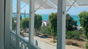 A view through a window with a white balcony railing, a patterned ceiling, and a clear view of the sea and surrounding greenery.
