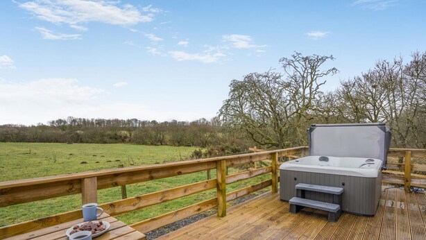 A wooden deck with a hot tub, picnic table with cups and chocolate, and a view of a green field and trees. - 2 Bed in Ochiltree (oc-w37517) (Ochiltree)