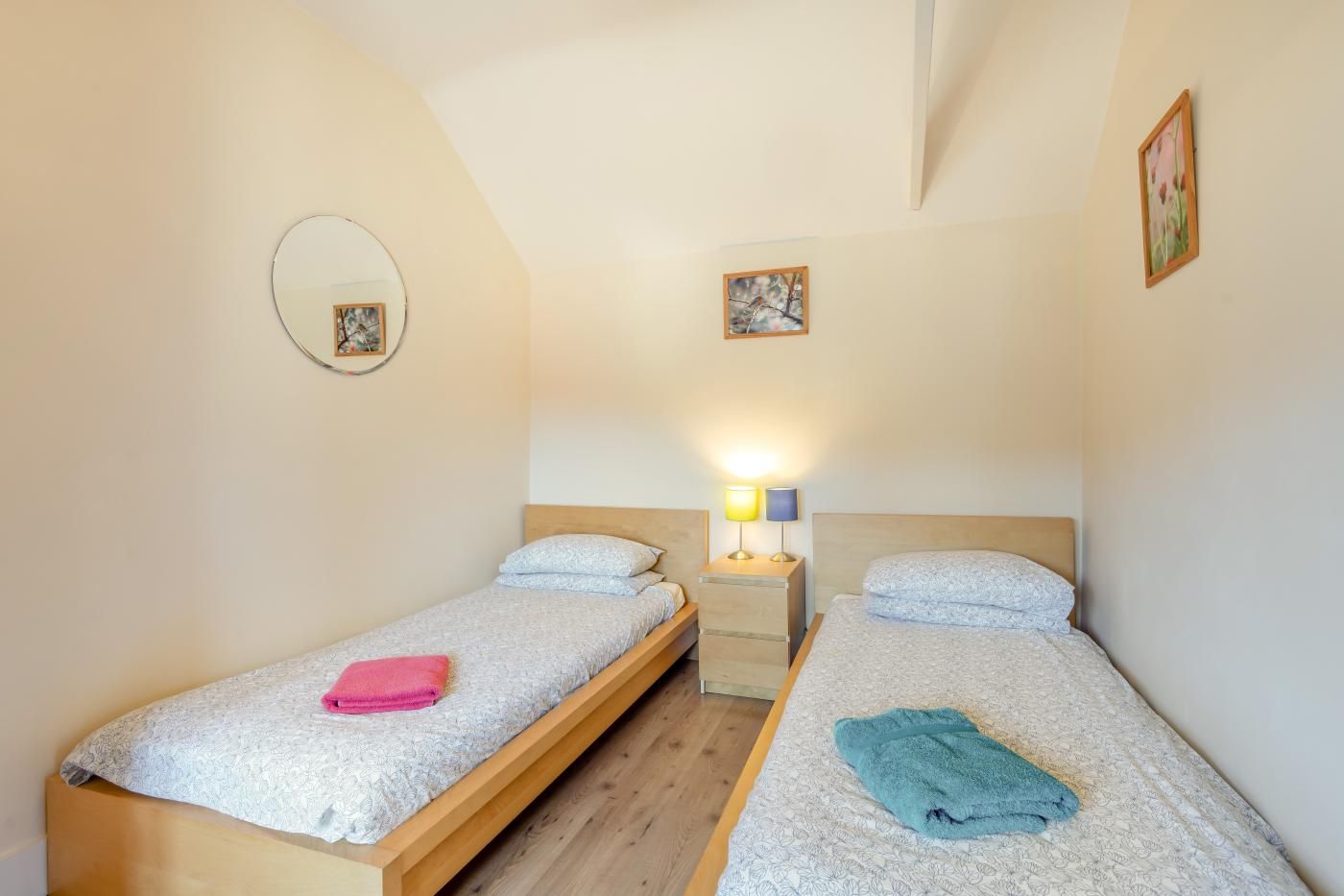 Une petite chambre agréable, dotée de deux lits simples, d’une table de chevet avec une lampe et d’un miroir fixé au mur.