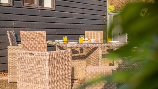 Ein Outdoor-Essetset aus Rattan mit Tisch und Stühlen, ein Glas Orangensaft und ein Teller mit Croissants, vor einem dunklen Holzbau platziert.