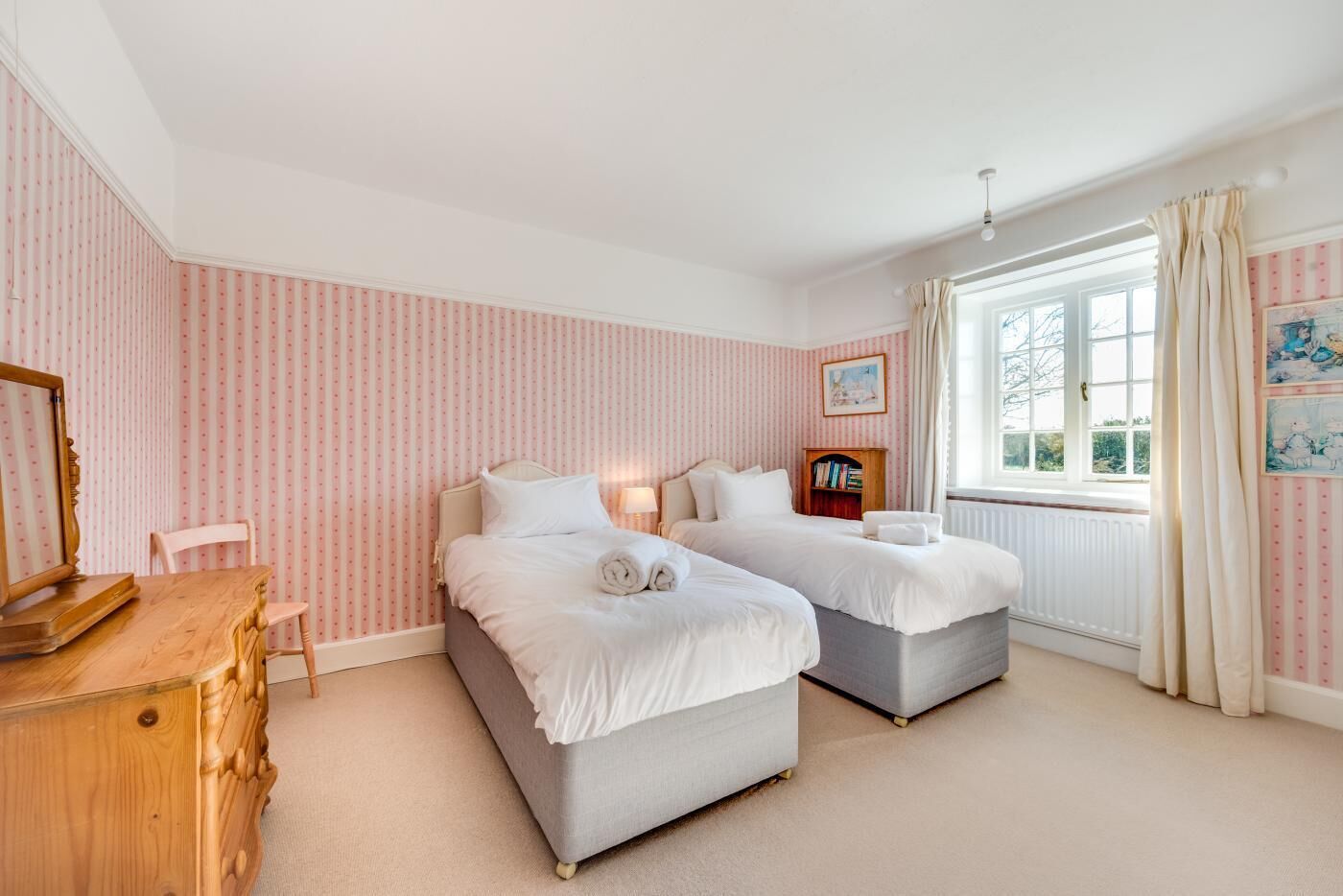 Een slaapkamer met twee bedden, een houten dressoir, een stoel, een boekenkast en een raam met gordijnen.