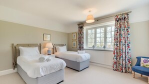 Une chambre avec deux lits, une fenêtre ornée de rideaux à motifs floraux, un fauteuil bleu et une lampe de chevet.
