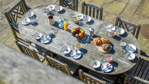 Une table en bois installée à l’extérieur pour un repas, avec des chaises, des assiettes, des couverts, un panier de fruits et du pain.