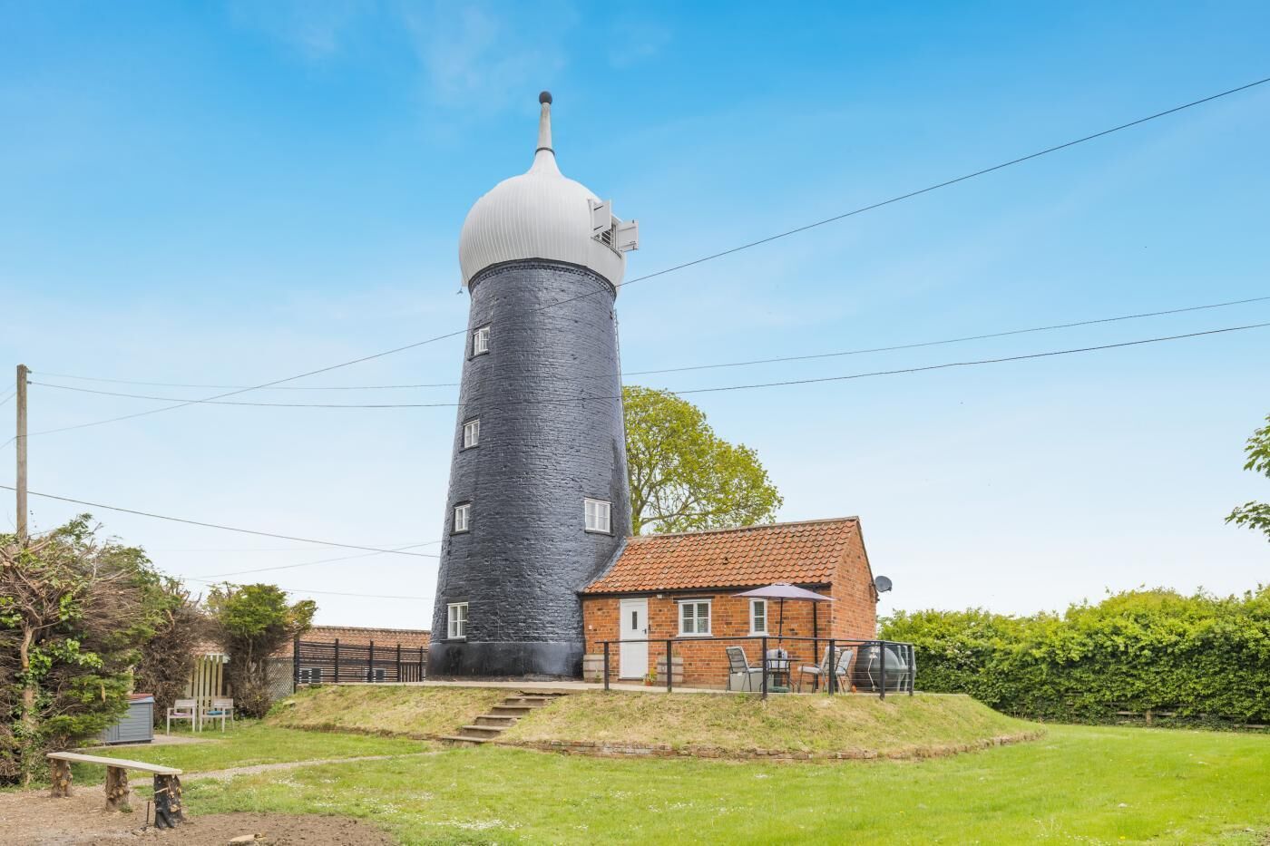 Un faro con una torre cilindrica e una cupola, un edificio in mattoni con tetto a tegole, e una zona erbosa con una panchina.
