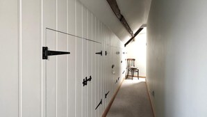 Een gang met witte paneelwanden en zwarte handgrepen, die leidt naar een kamer met een houten stoel en een raam.